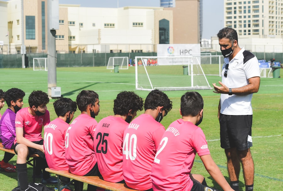 A man in a pink shirt giving instructions to a group of young soccer players on the field.
